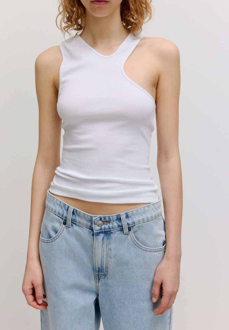 Witte mouwloze top met een asymmetrische halslijn en een gladde textuur, gecombineerd met lichtblauwe denimbroek met lichte vervaging en zakken.
