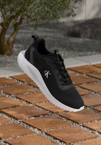 Černé sportovní tenisky s bílou podrážkou a logem 'cK', plovoucí nad venkovním povrchem z dřevěných dlaždic a štěrku s rostlinami v pozadí.