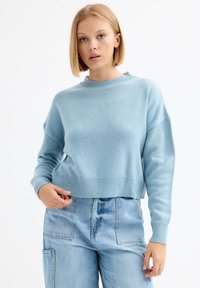 Suéter de punto azul claro con cuello redondo, hombros caídos y puños acanalados. Combinado con pantalones de mezclilla claros de cintura alta.