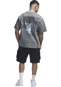 Homme portant un T-shirt gris oversized avec le texte « INTROSPECT » et un graphisme, un short cargo noir, des chaussettes blanches et des baskets blanches, debout de dos.