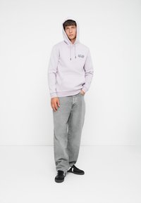 Light purple hoodie with front pocket, drawstring hood, and black logo. Paired with loose grey jeans and black sneakers.