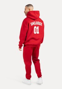 Červená souprava mikiny a tepláků. Mikina má kapuci a na zádech je bílý nápis "SMILODOX 01". Materiál vypadá měkký a strukturovaný.