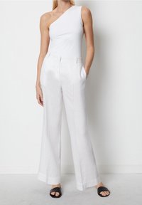 Bodysuit blanc à une épaule associé à un pantalon en lin blanc ample. Modèle portant des sandales à glissière noires, avec des poches avant visibles.