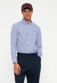 Gai zili un balti svītraini krekli ar garām piedurknēm un apkakli, saskaņoti ar tumši ziliem biksēm un burgundijas krāsas cepuri.