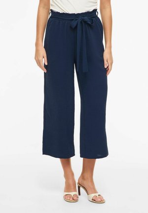Femme portant un pantalon large cropped bleu marine avec ceinture nouée à la taille, haut blanc et sandales à talons ouvertes beige, debout sur un fond blanc.