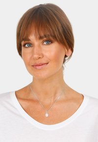Silberne Halskette mit einem ovalen, rosafarbenen Anhänger, getragen über einem weißen Hemd, mit einer glatten Textur und minimalistischem Design.