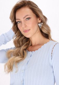 Cárdigan de punto azul claro con escote ondulado, canalé vertical y detalles de botones de perlas, con acentos transparentes en las mangas.