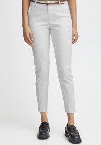 Une personne se tient debout, portant un pantalon gris clair ajusté, un haut blanc et des mocassins noirs. Elle a les mains le long du corps contre un fond uni.