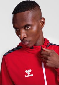 Rote Reißverschlussjacke aus strukturiertem Stoff. Mit schwarzen geometrischen Akzenten an den Schultern und einem weißen Logo auf der Brust.