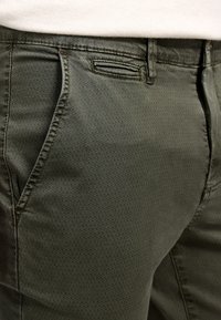 Pantalones verde oliva hechos de un tejido texturizado, con un patrón geométrico, bolsillos tradicionales y un detalle de cinturilla cosido.