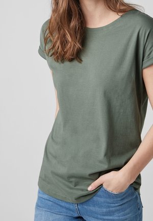 Kvinde iført en kortærmet olivengrøn t-shirt og lyseblå jeans, med den ene hånd afslappet i lommen, mod en ensfarvet baggrund.