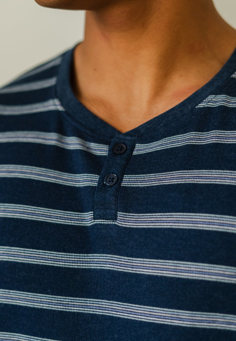 Marineblauwe gestreepte henley-shirt met een zachte textuur, twee donkere knopen en een V-halsontwerp met afwisselend witte en lichtgrijze strepen.