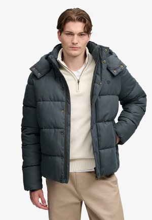Ung mand iført en mørkegrå dunjakke over en cremefarvet kvart lynlås sweater og beige bukser, stående med den ene hånd på hoften.