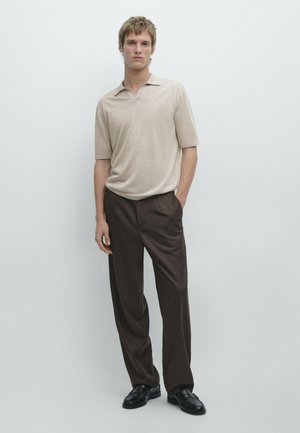 Young man standing against a plain wall wearing a beige short-sleeve polo shirt, brown wide-leg trousers, and black shoes.