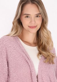 Texturerad, ljusrosa cardigan med öppen front, bärs över en vit topp. Mjuk tyg, lös passform och avslappnad design. Naturligt hår som ramar in ansiktet.