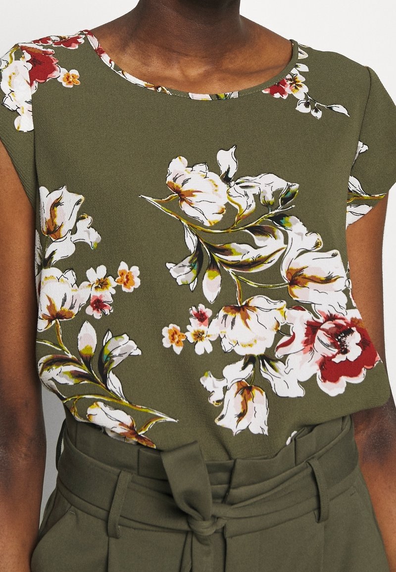 Gros plan d'une personne portant un haut floral vert olive avec des fleurs blanches et rouges, associé à un pantalon taille haute vert olive noué à la taille.