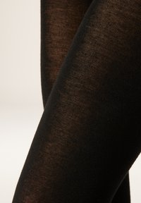 Svarta, genomskinliga strumpbyxor med en slät textur, som har ett fint stickat mönster. Närbilden visar en subtil lyster och benkonturer.