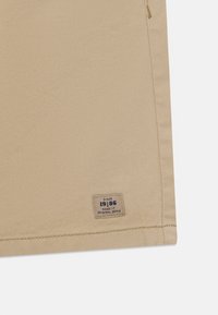 Bežinis audinys su lygiu paviršiumi, kuriame pritvirtinta stačiakampė etiketė su užrašu „SINCE 19186 NAME IT ORIGINAL DENIM“ mėlyna spalva.