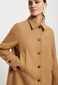 Kaban od mješavine deva sa strukturiranim ovratnikom, prednjim dugmadima i velikim bočnim džepovima. Glatka tekstura s toplim, neutralnim tonom.