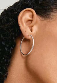 Orecchino a cerchio in argento esposto in un orecchio forato. L'orecchino è circolare, caratterizzato da una superficie liscia e riflettente e un design minimalista.
