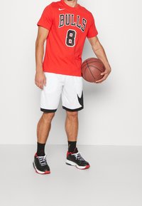 Roten Bullen T-Shirt mit schwarzer Schrift und der Nummer 8, kombiniert mit weißen Shorts mit schwarzen Akzenten und schwarzen Sportschuhen mit roten Details.