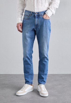 Mężczyzna ubrany w jasnoniebieskie jeansy, białe sneakersy i białą, długorękawową koszulę zapinaną na guziki, z jedną ręką w kieszeni.