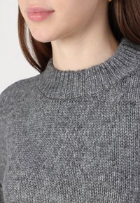Šedý pletený svetr s ribbovaným výstřihem a texturovanou tkaninou. Detailní záběr se zaměřuje na oblast krku a zdobné švy.