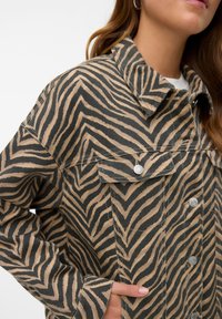 Prindi zebra triibudega jakk, beež ja must, millel on krae, nööbid ja ees olevad taskud. Materjal näeb välja tekstuurne ja vastupidav.