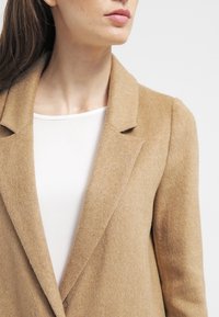 Kabát z kašmírové vlny s kamelovým podílem, se strukturovaným střihem, klopami s výřezem a jemnou texturou. Nošený přes bílý top, zdůrazňující čistý design.