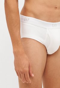 Slip in cotone bianco con logo GAP sull'elastico in vita. Presenta un design aderente con una superficie liscia e aperture laterali.