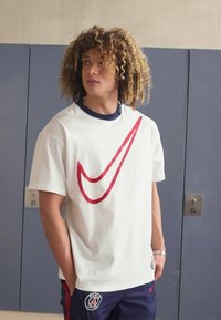 Camiseta blanca de algodón con un gran diseño del logo de Nike en rojo. La camiseta tiene un cuello redondo azul marino y se combina con pantalones cortos azul marino.
