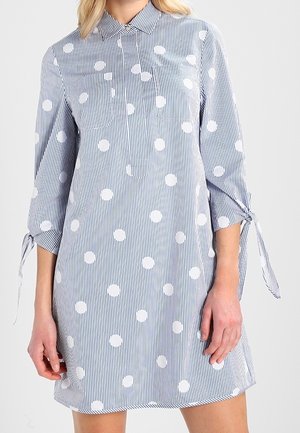 Kobieta ubrana w sukienkę w pionowe niebiesko-białe paski z dużymi białymi kropkami i rękawami o długości 3/4 związanymi przy mankietach.