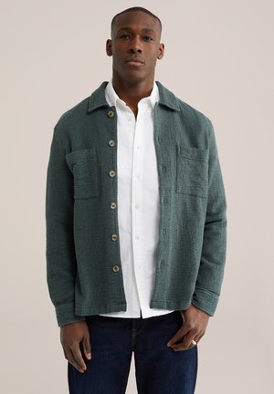 Mężczyzna ubrany w ciemnozieloną, teksturowaną kurtkę na guziki, pod którą ma białą koszulę, oraz ciemnoniebieskie dżinsy, stoi na tle jasnej, jednolitej ściany.