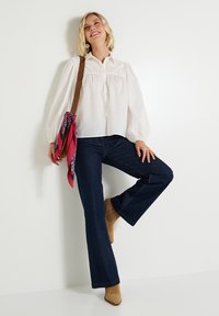 Blusa blanca con mangas abullonadas y un canesú bordado, combinada con jeans acampanados de color azul oscuro. Botines de ante marrón y un pañuelo rosa como acento.