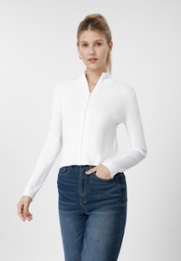 Camisola branca com fecho de correr canelada e design cropped, com mangas compridas e gola dobrada, combinada com jeans de ganga azul escuro.