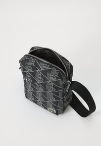 Borsa a tracolla a pattern nero e grigio con zip argentata, ampia tracolla nera e piccolo logo di coccodrillo verde nell'angolo anteriore.