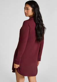 Bordeauxrotes Langarmkleid mit Kragen, lockerem Schnitt, seitlichen Schlitzen und glatter Stoffstruktur. Minimalistische Designelemente.