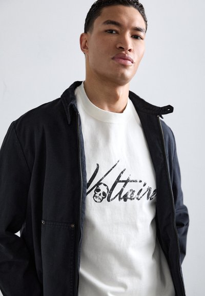 Chaqueta negra con cremallera sobre una camiseta de cuello redondo blanca, que presenta un diseño gráfico negro con una calavera y el texto "Voltaire". Corte casual.