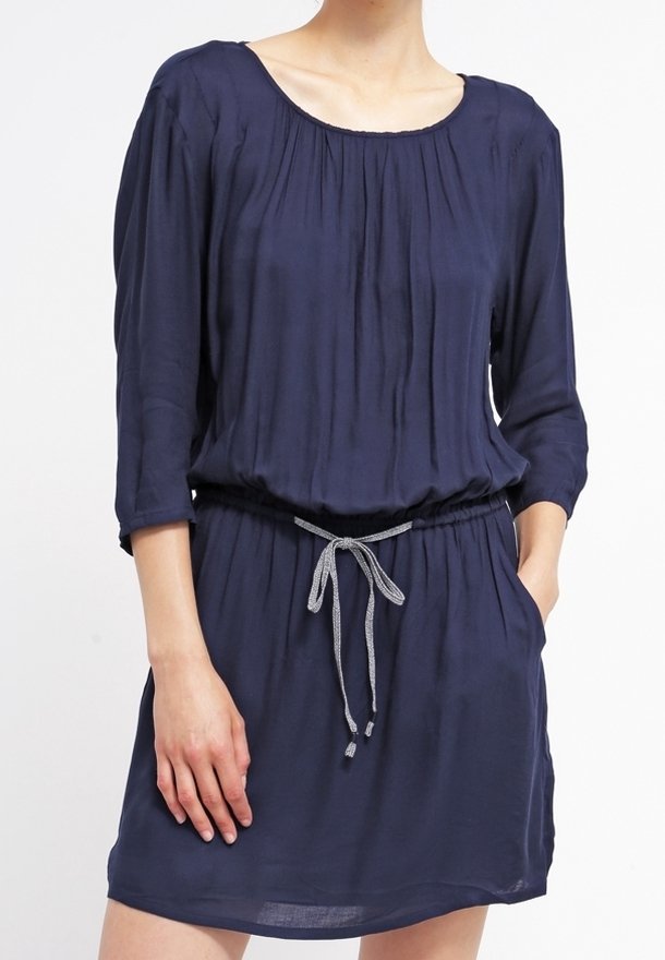 Femme portant une robe bleu marine à manches longues avec un col froncé et un cordon gris noué à la taille, une main dans la poche.