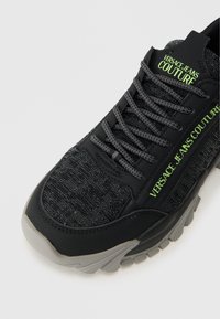 Čierne tenisky s textúrovaným textilným zvrškom, šedou podrážkou a zeleným logom "VERSACE JEANS COUTURE". Obsahujú šnúrky a spevnenú špičku.