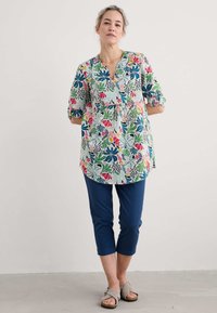 Kwiecowa tunika z lekkiego materiału, z dekoltem w serek i rękawami 3/4. Połączona z granatowymi spodniami capri i jasnoszarymi sandałami.