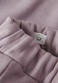 Un tissu violet doux et léger présente une taille froncée avec une fermeture à bouton et des détails texturés, idéal pour le confort et la facilité de port.