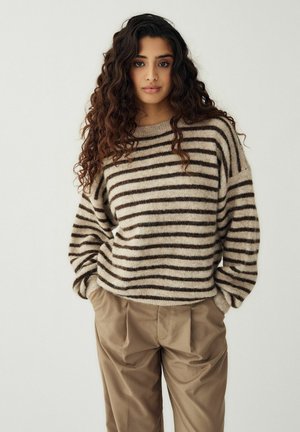 Hosszú, göndör hajú nő bézs és barna csíkos pulóvert, valamint bő, khaki nadrágot viselve, zsebeiben tartott kezekkel áll.