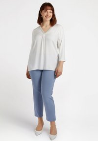 Weiße Bluse mit V-Ausschnitt und strukturiertem Saum, kombiniert mit hellblauen figurbetonten Hosen. Hat halbe Ärmel und einen lockeren Schnitt.