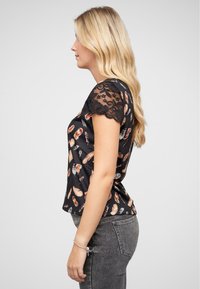 Haut noir avec un motif de plumes dans des tons de marron et d'orange, avec des manches courtes en dentelle et une coupe ajustée. Porté avec un jean gris.