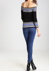 Frau mit schwarz-weiß gestreiftem, schulterfreiem Pullover, blauen Skinny Jeans und schwarzen hochhackigen Stiefeln, die vor weißem Hintergrund steht.