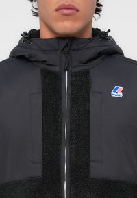 Veste noire avec un panneau en polaire texturé noir à l'avant, fermeture éclair, capuche ajustable et un logo K-WAY sur le côté supérieur gauche.
