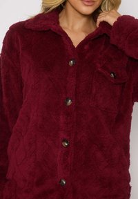 Giacca camicia in pile bordeaux con motivo a diamante a trama, bottoni frontali e una tasca sul petto. Tessuto morbido e accogliente.