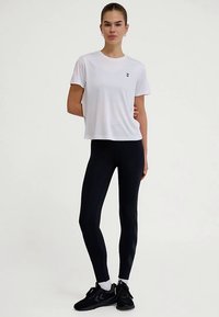 Biała koszulka z krótkim rękawem z logo na piersi, zestawiona z czarnymi legginsami o fakturowanym wzorze oraz czarnymi butami sportowymi.