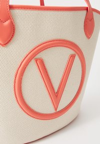 Crème canvas draagtas met een groot koraal logo met een "V"-ontwerp en leren accenten, waaronder handvatten en rand.
